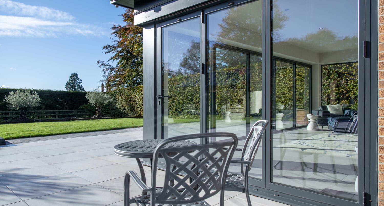 A small metal table and two chairs sit on a patio outside bifold doors, overlooking a sunny garden with a green lawn and trees.