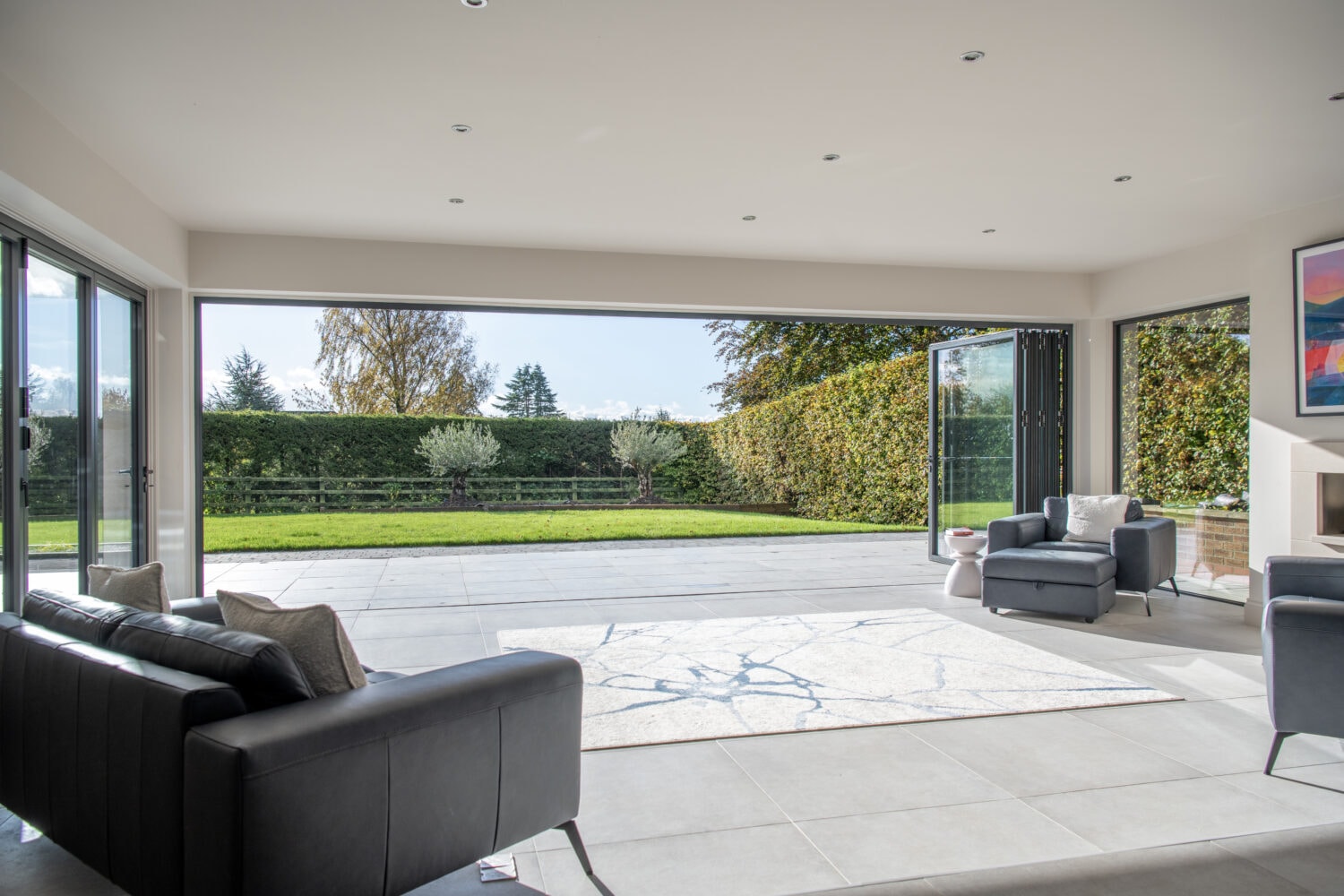 Modern living room with large bifold doors fully opened to a spacious patio and green lawn, allowing natural light to flood the space. There are black sofas, a gray armchair, and a light-colored rug inside.