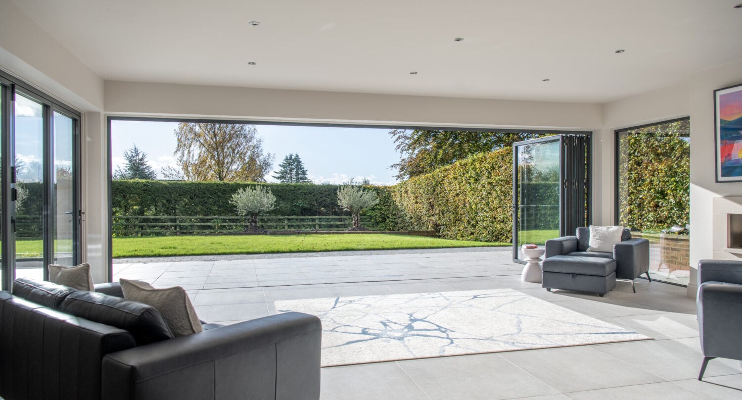 Modern living room with large bifold doors fully opened to a spacious patio and green lawn, allowing natural light to flood the space. There are black sofas, a gray armchair, and a light-colored rug inside.