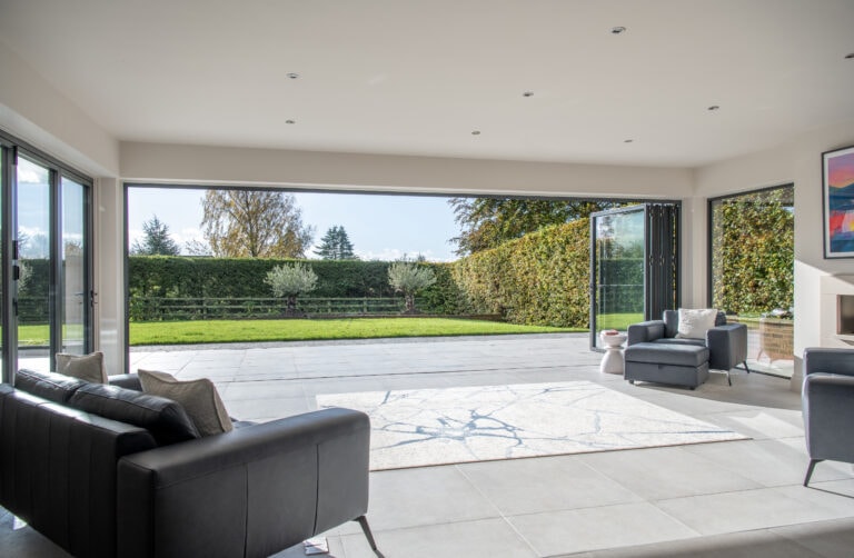 Modern living room with large bifold doors fully opened to a spacious patio and green lawn, allowing natural light to flood the space. There are black sofas, a gray armchair, and a light-colored rug inside.