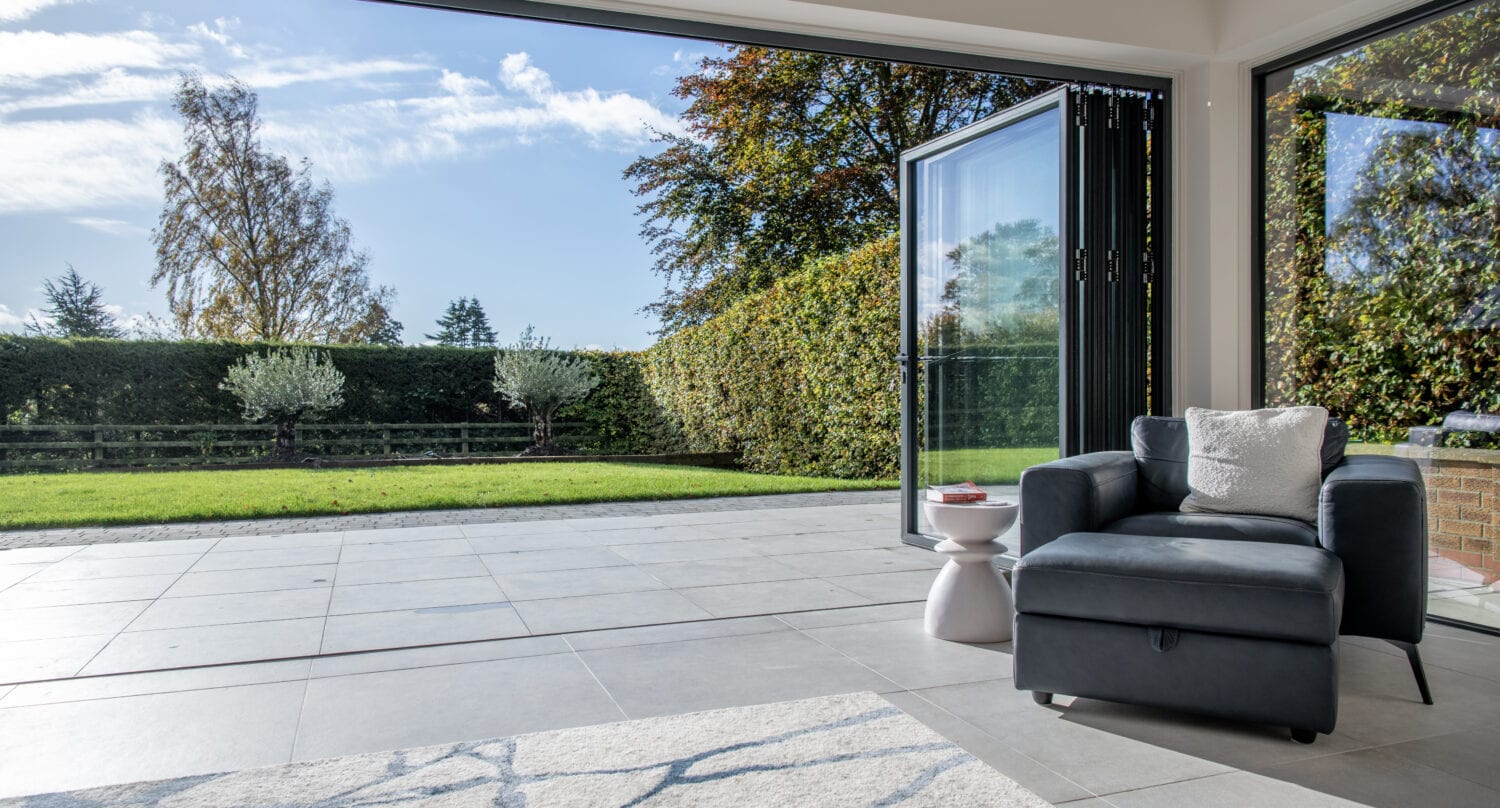 A modern living space with large bifold doors open to a green garden; a dark armchair and small white side table sit on tiled flooring near a gray patterned rug. Trees and a blue sky are visible outside.