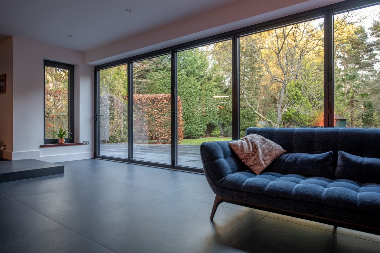 Modern living room with large floor-to-ceiling sliding doors overlooking a patio and lush green garden. A dark blue sofa with cushions sits on gray tile flooring near the windows.