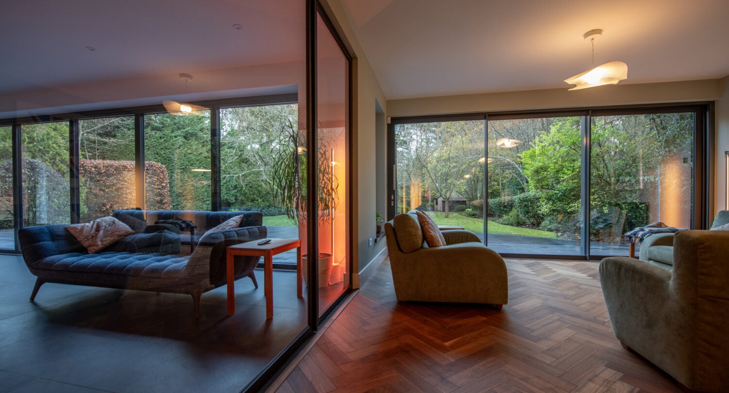 Modern living room with wood flooring, large floor-to-ceiling windows, cozy sofas, and views of a green garden. Bespoke glass solutions include a glass wall dividing two sunlit seating areas, seamlessly connecting to the outdoors.
