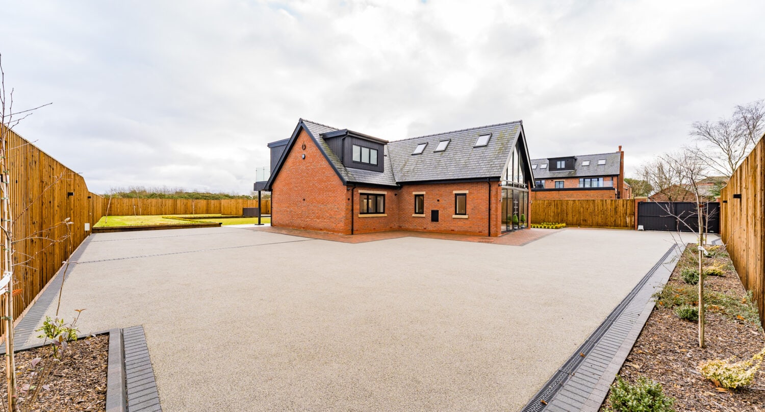 A modern brick house with large windows, bespoke glass solutions, and a black roof sits in the center of a spacious, paved driveway, enclosed by wooden fencing and landscaped garden beds under a cloudy sky.