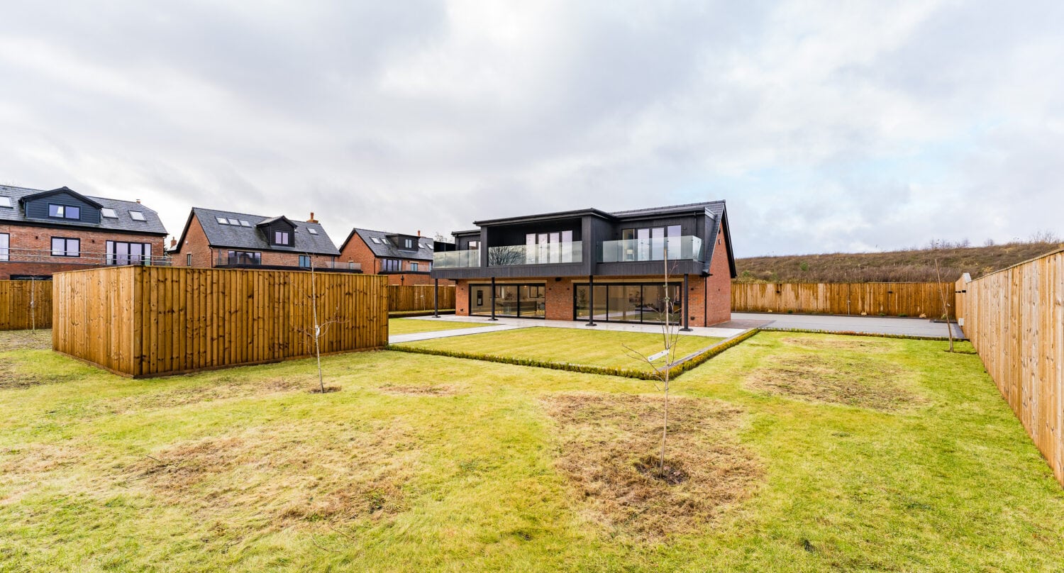 Modern two-story house with large windows, sliding doors, and a balcony, surrounded by a spacious fenced backyard with patches of grass. Other similar houses are visible in the background under a cloudy sky.