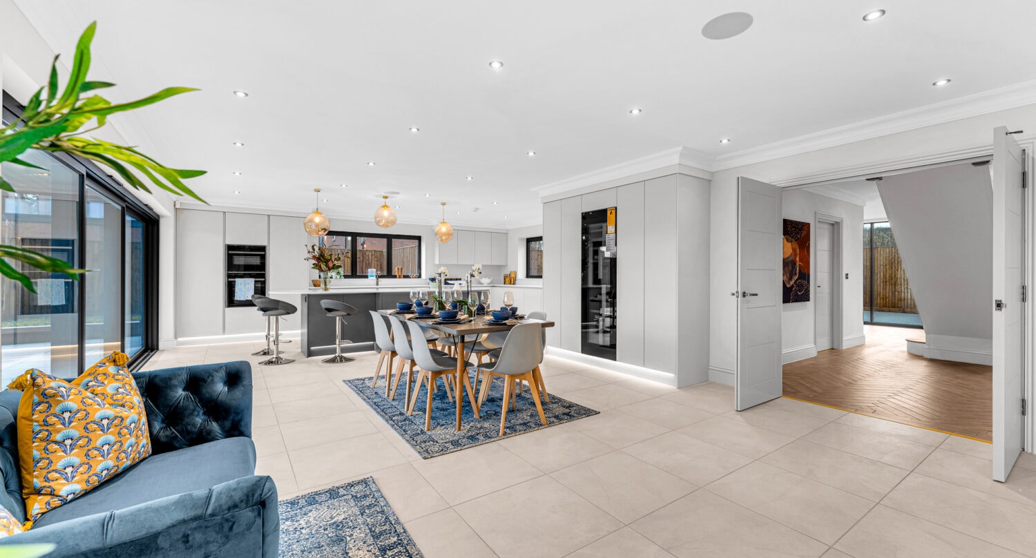 Open-plan, modern living and dining space with white walls, tiled floors, a blue sofa with yellow cushions, a dining table set for six, a kitchen with bar stools, large windows, sliding doors, and recessed ceiling lights.