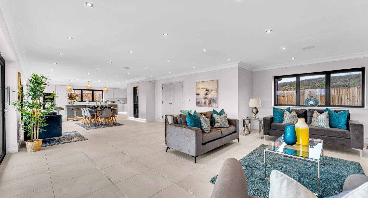 Spacious modern open-plan living area with gray sofas, blue and yellow accents, large windows, tiled floors, a dining area, and a kitchen in the background. Sliding doors and green plants complement the contemporary artwork decorating the room.