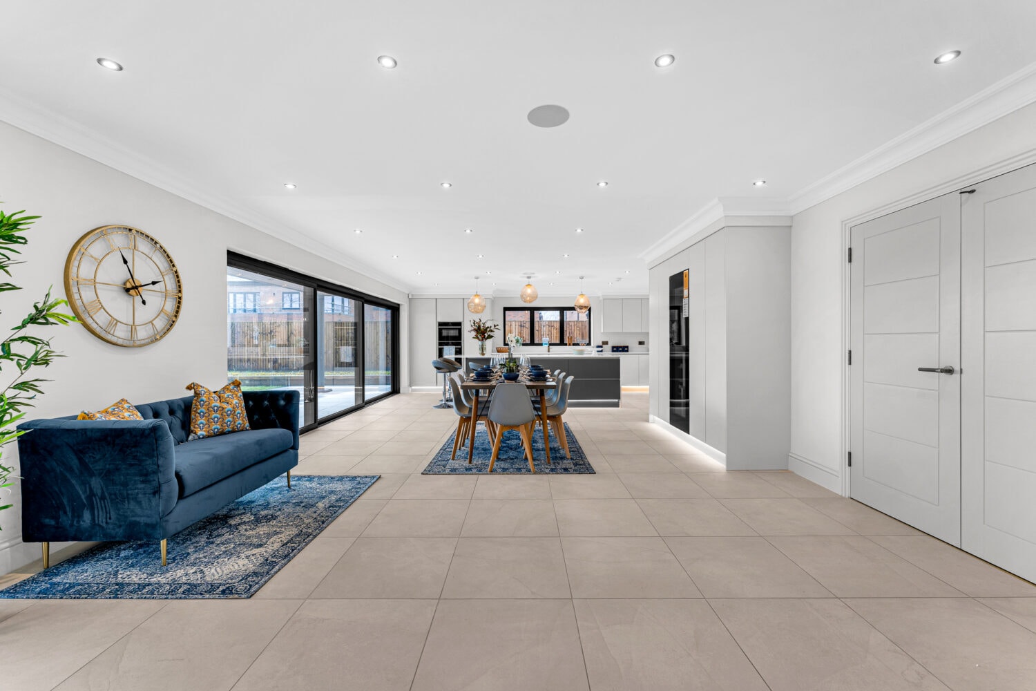 Spacious modern open-plan living and dining area with a blue sofa, dining table, large kitchen island, tiled floor, contemporary decor, bifold doors, large windows, and plenty of natural light.