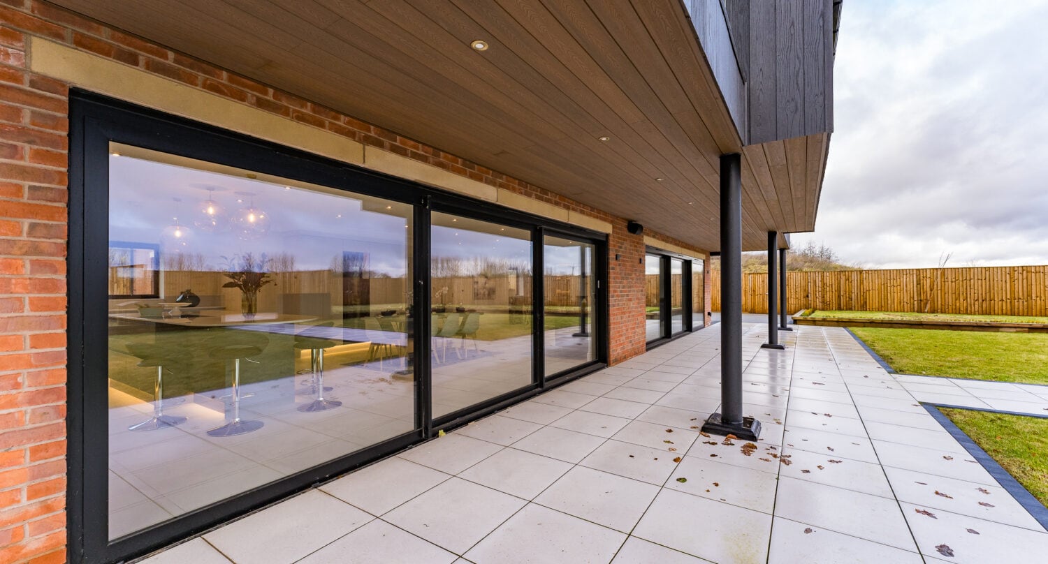 A modern house with large sliding doors opens onto a spacious tiled patio. The yard features green grass, a wooden fence, and overcast skies are visible in the background, highlighted by bespoke glass solutions.