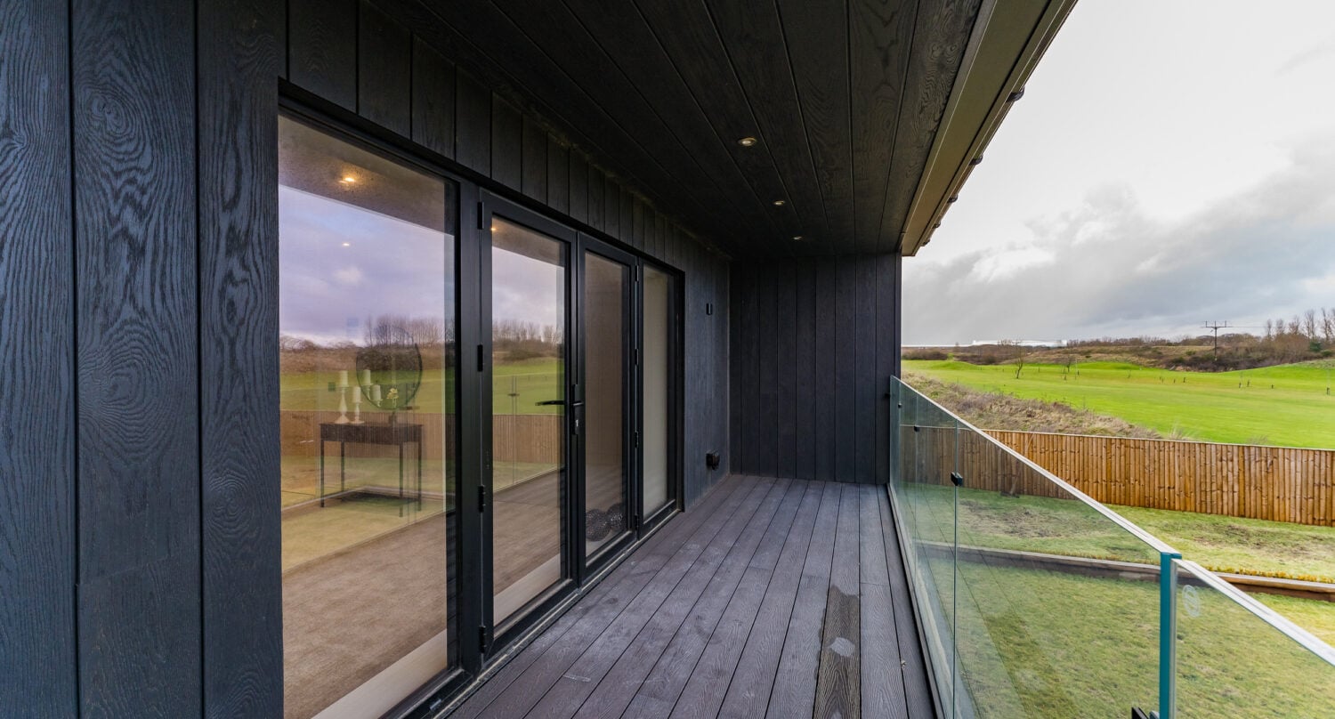 A modern balcony with dark wood paneling, glass railing, and recessed lighting overlooks a green field and distant trees on a cloudy day. Reflections of the interior are visible in the sliding glass doors.