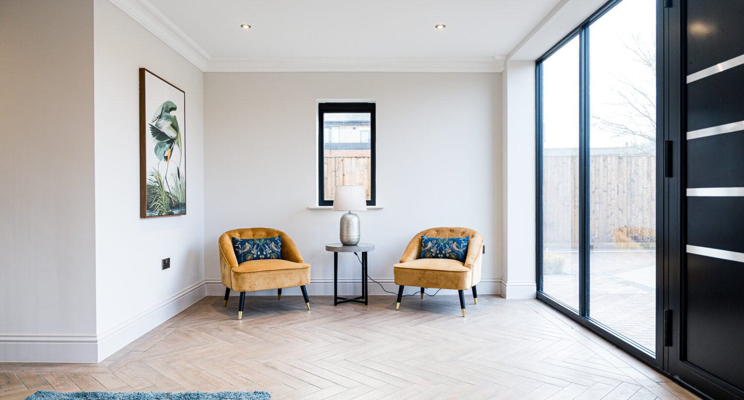 A modern, minimalist room with two mustard yellow chairs, a small black table with a decorative vase, framed bird print, herringbone flooring, and large bespoke glass sliding doors letting in natural light.