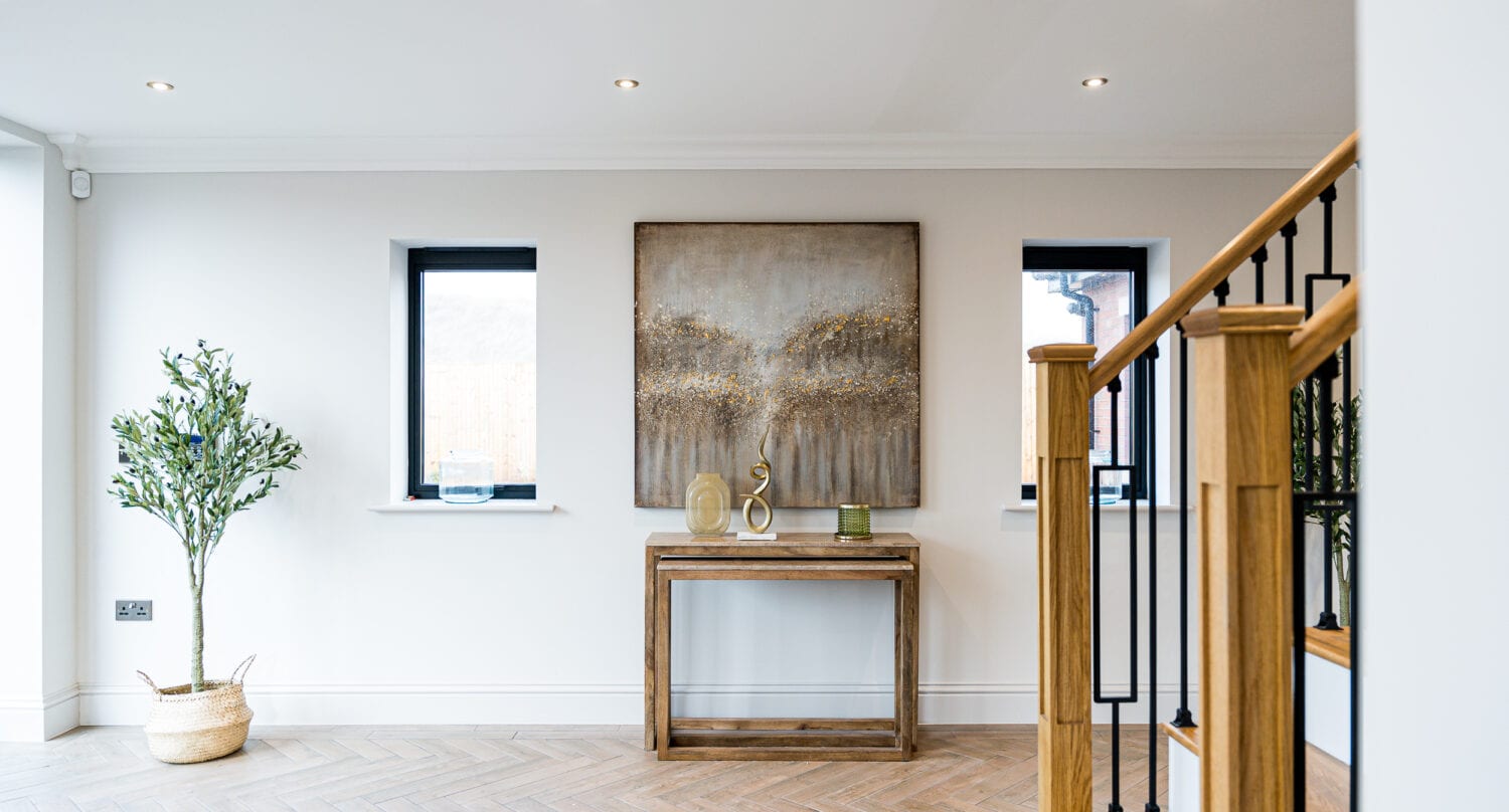 Modern hallway with wooden stairs, a potted plant, blue rug, and a slim wooden table displaying decor items beneath a large textured artwork, with bespoke glass solutions seen in the black-framed windows enhancing natural light.