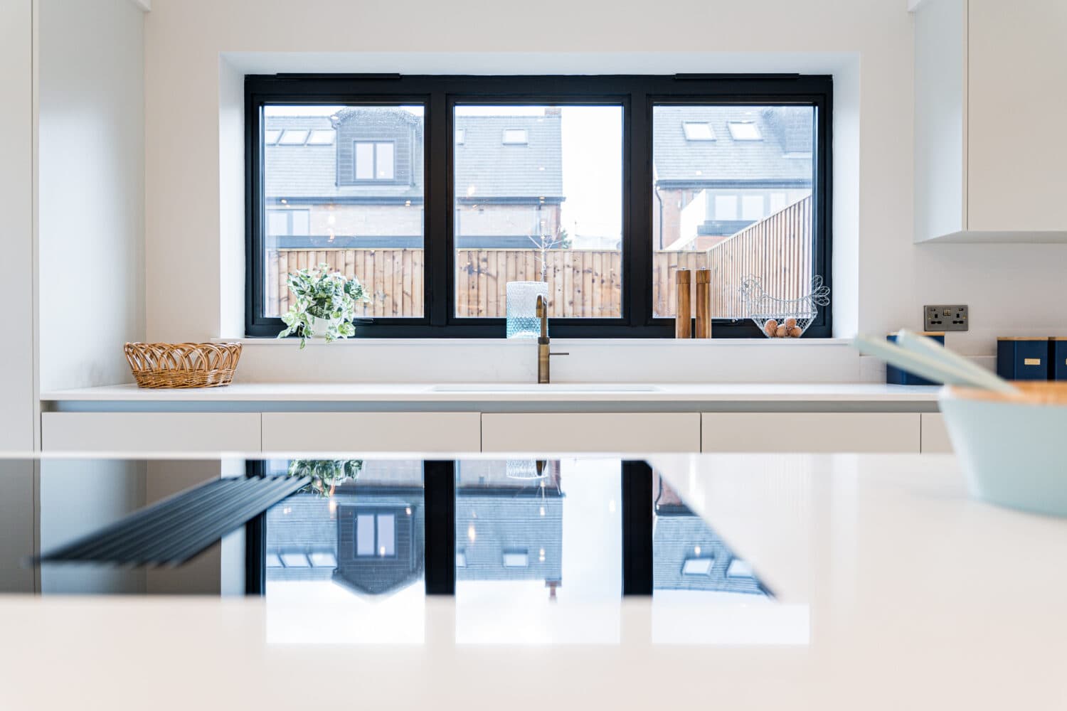 Modern kitchen with a white countertop, sink under a large black-framed window, basket and plant to the left, wooden utensils to the right, and reflections of houses or sliding doors outside visible on the counter.