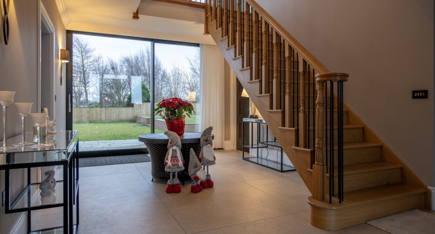 A modern entryway with a wooden staircase, glass table with a poinsettia plant, two decorative gnomes, and large windows featuring bespoke glass solutions that showcase a grassy yard with trees outside.