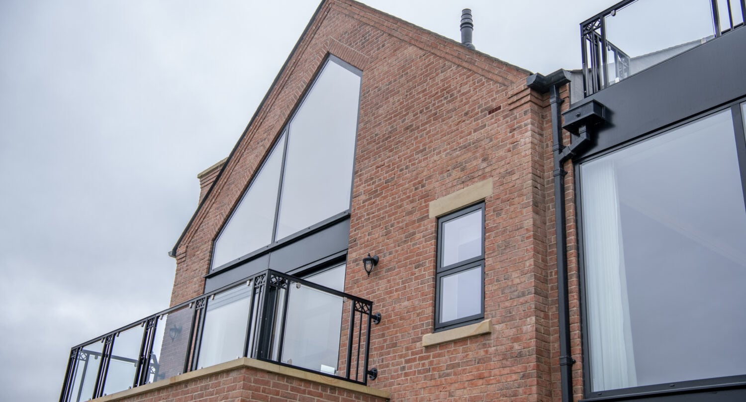 Modern brick house with large triangular and rectangular windows, black frames, glass balcony railing, and sleek sliding doors, set against a cloudy sky.