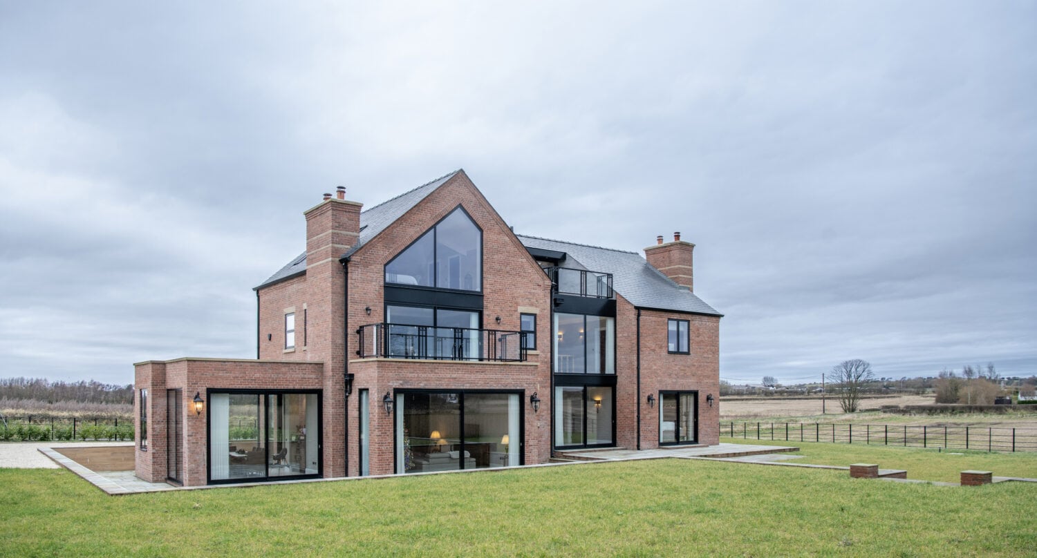 A modern two-story brick house with large glass windows, sliding doors, and balconies sits on a spacious green lawn, surrounded by open fields under a cloudy sky.