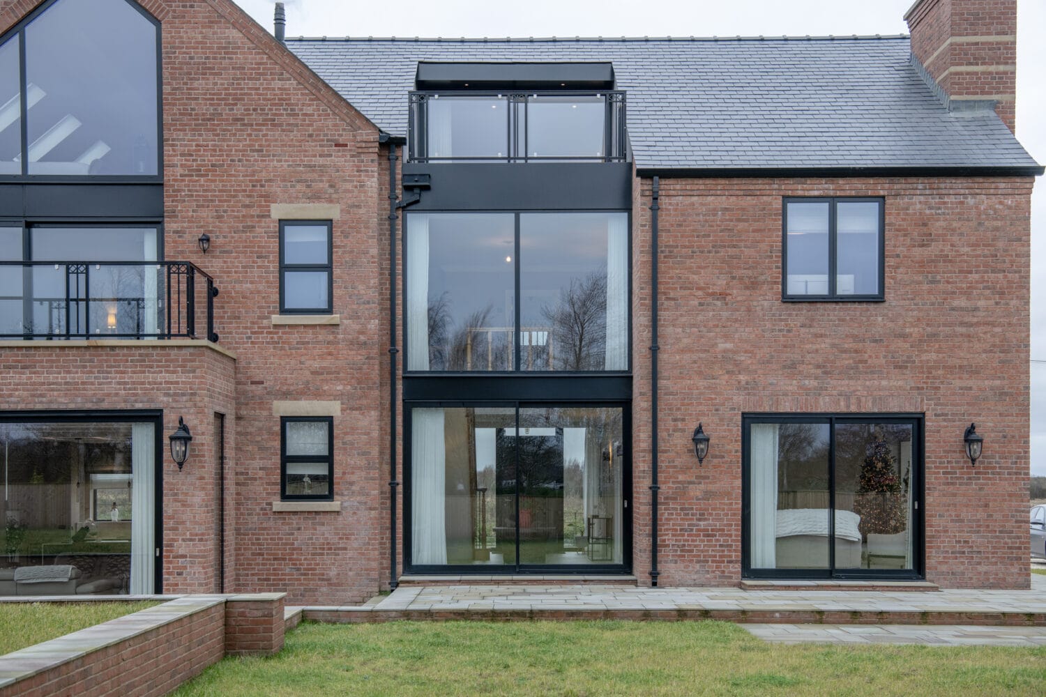 Modern red brick house with large glass windows and bifold doors, a small balcony on the left, and a patio with stone paving in front. The lawn is green, and trees are visible through the windows.