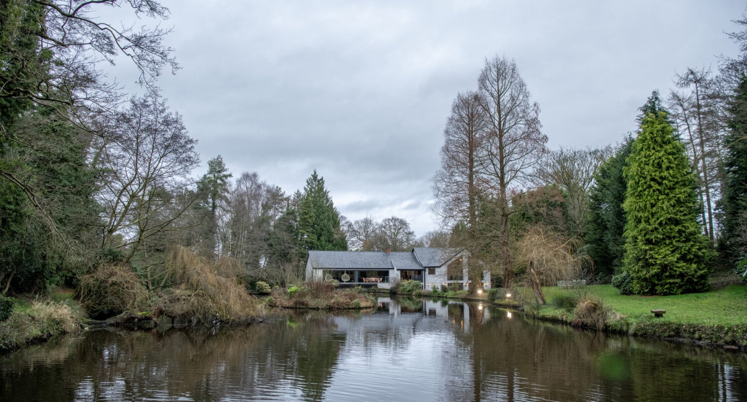 A small house with glass roofing sits by the edge of a calm pond, surrounded by trees and greenery under a cloudy sky. The water reflects the house and trees, creating a peaceful, natural scene.