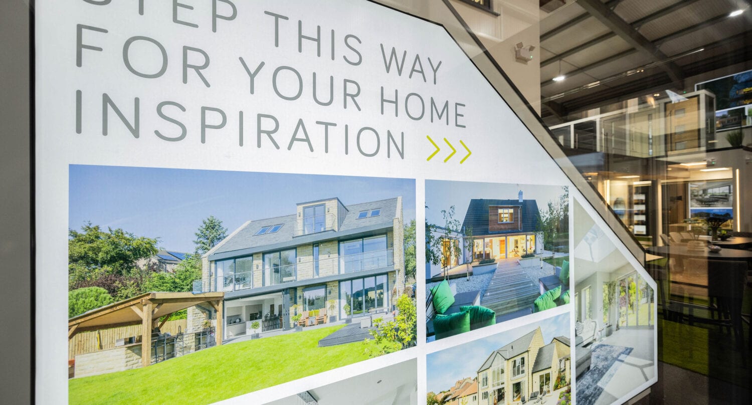 Large sign with “Step This Way For Your Home Inspiration” above four photos of modern homes, displayed inside a showroom featuring bespoke glass solutions and other home design elements in the background.