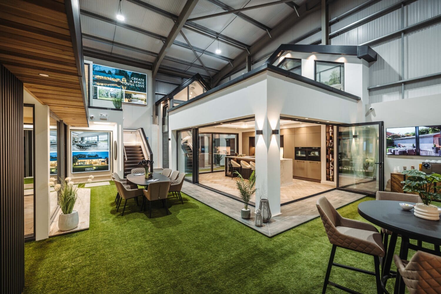 A modern showroom with artificial grass floor displays stylish indoor and outdoor living spaces, featuring seating areas, plants, and bespoke glass solutions such as bifold doors under high metal ceilings with bright lighting.