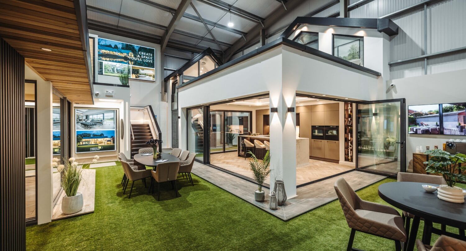 A modern showroom with artificial grass floor displays stylish indoor and outdoor living spaces, featuring seating areas, plants, and bespoke glass solutions such as bifold doors under high metal ceilings with bright lighting.