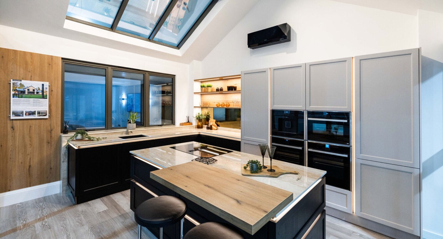 Modern kitchen with skylight, large windows, sleek black and wood cabinets, built-in ovens, island with wooden countertop, two round black stools, minimalist decor, potted plants, shelves, and bespoke glass solutions like sliding doors for a seamless look.