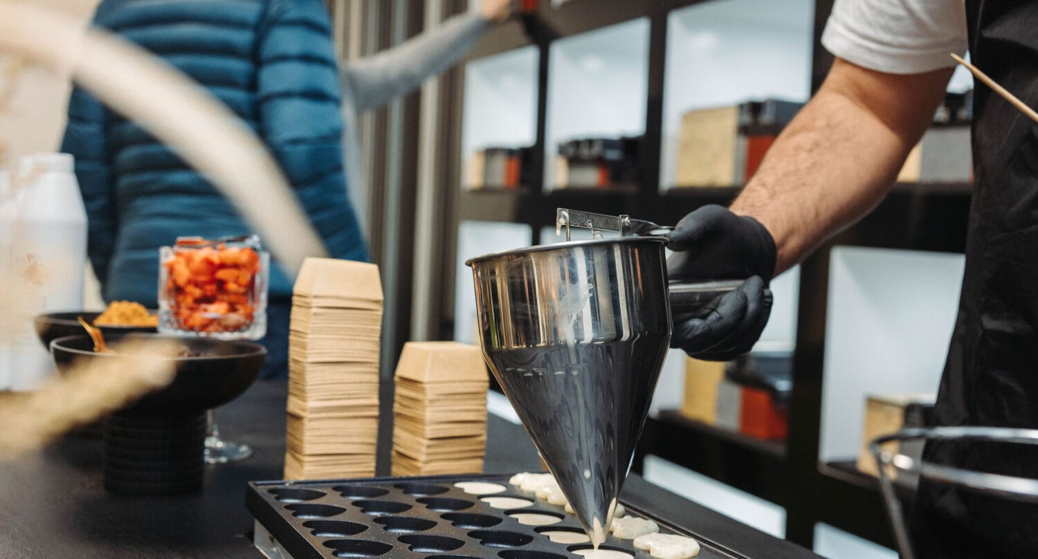 A person wearing black gloves pours batter from a metal funnel into a baking tray with circular molds, while shelves filled with various jars are reflected in bespoke glass solutions, adding elegance to the kitchen scene.