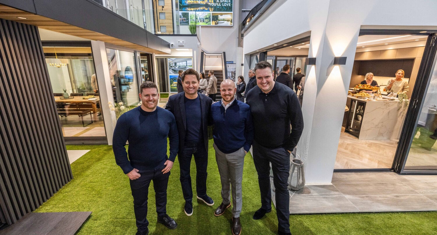 Four men stand smiling on artificial grass inside a modern home showroom with large glass windows and sliding doors. Behind them, people gather in the kitchen area, surrounded by stylish interiors and bespoke glass solutions.