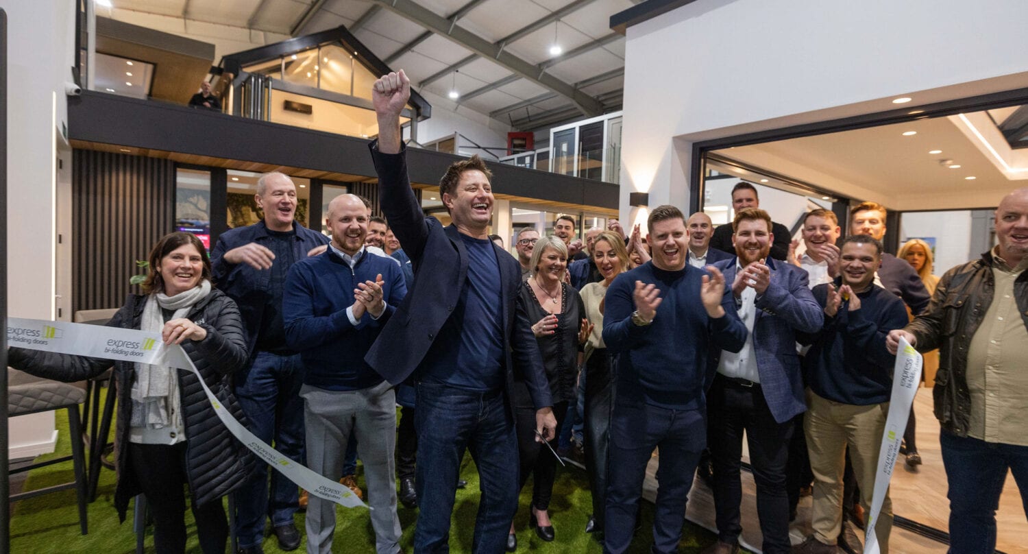 A group of people cheer and clap as a man in the center raises his fist in celebration at a ribbon-cutting ceremony inside a modern indoor space, where modular homes boast stylish bifold doors in the background.