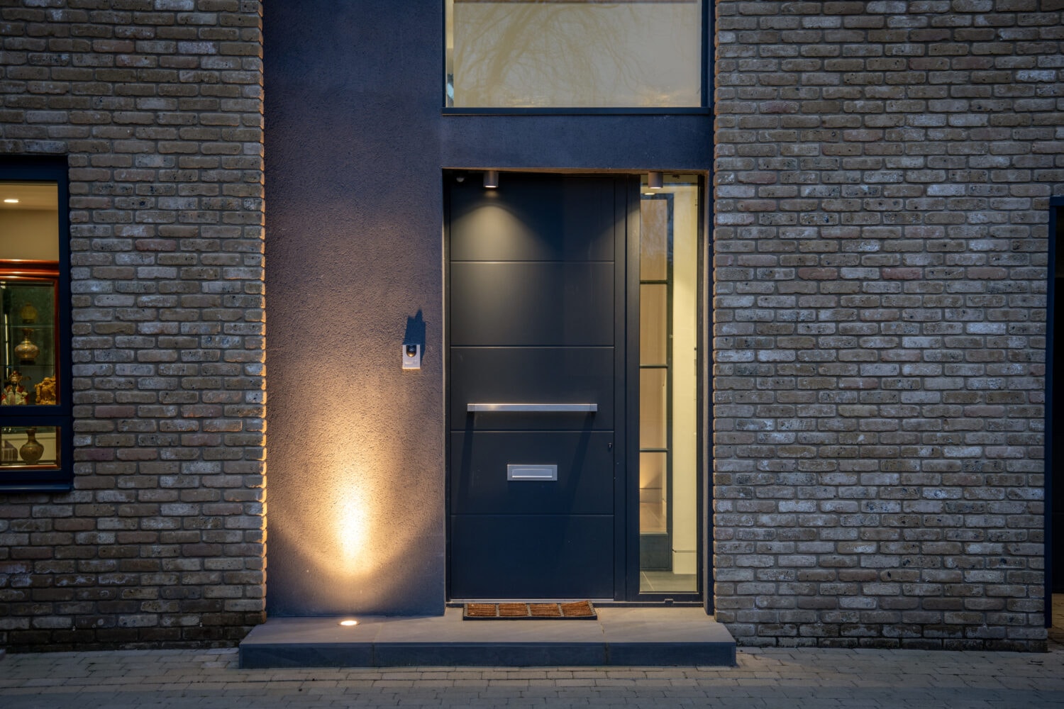 Modern front door with a sleek black finish, surrounded by light brown brick walls and illuminated by a spotlight from below. A narrow window beside the door showcases our bespoke glass solutions, while a small welcome mat sits on the doorstep.