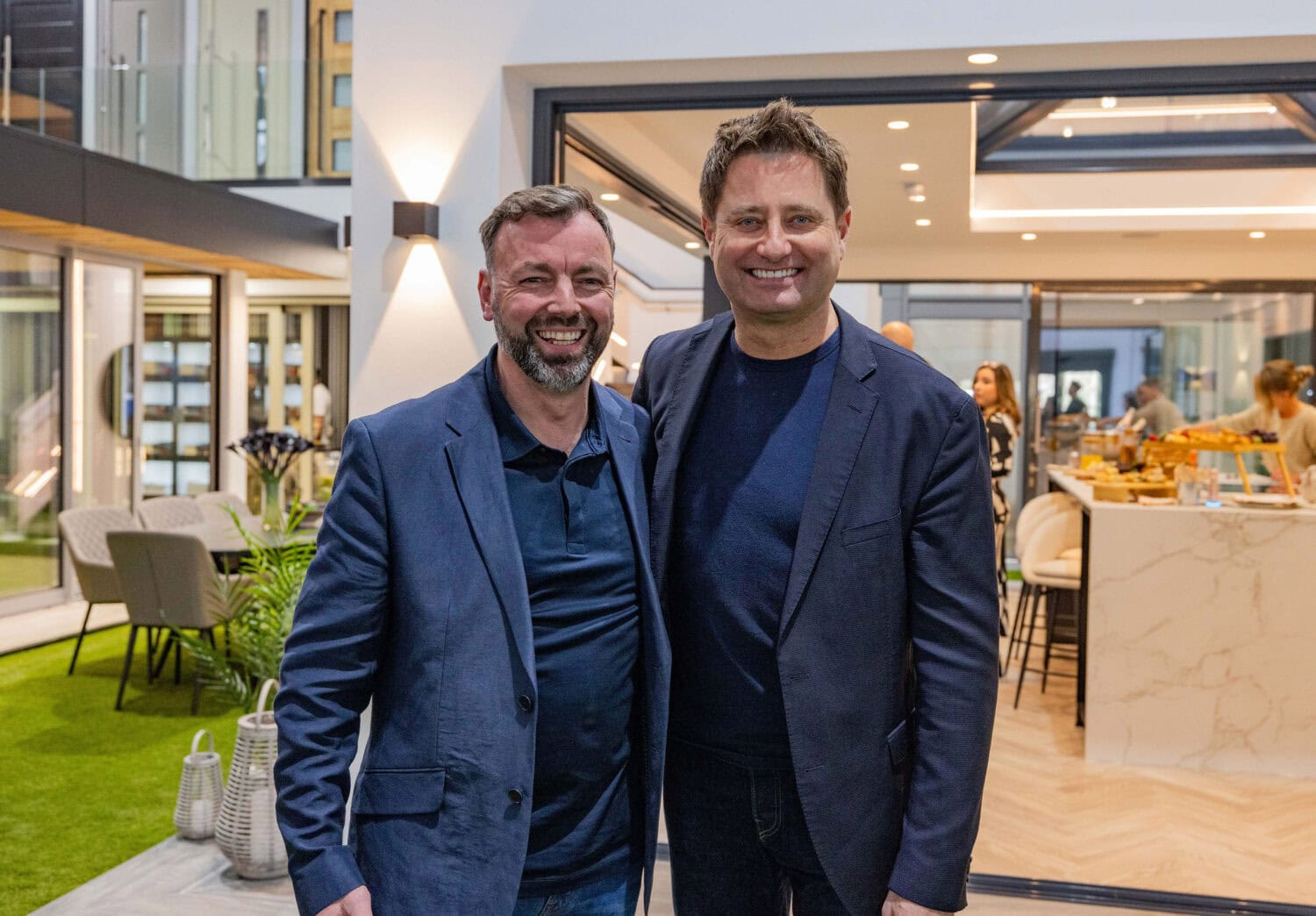 Two men wearing dark blazers stand smiling together in a modern indoor-outdoor space with large windows, bifold doors, lush plants, and people mingling in the background.