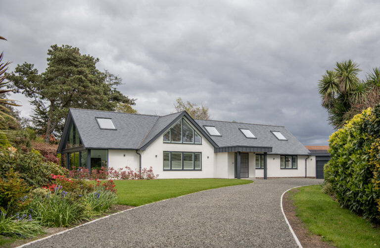 Modern single-story house with large windows, sleek glass roofing, and a dark gray sloped roof, surrounded by a well-kept lawn, colorful flowers, and trees under a cloudy sky. A gravel driveway leads up to the entrance.
