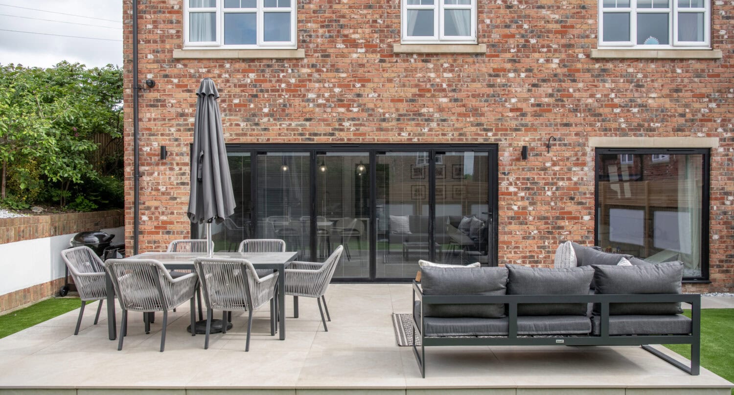 A modern brick house with bespoke glass solutions features large bifold doors that open onto a patio with a gray outdoor sofa, dining table, chairs, and a closed umbrella. The patio is surrounded by artificial grass and greenery.