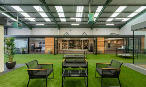 Modern indoor space with artificial grass flooring, black metal and mesh chairs around a glass table featuring bespoke glass solutions, small plants, and large windows showing office areas in the background. Industrial ceiling with bright overhead lights.