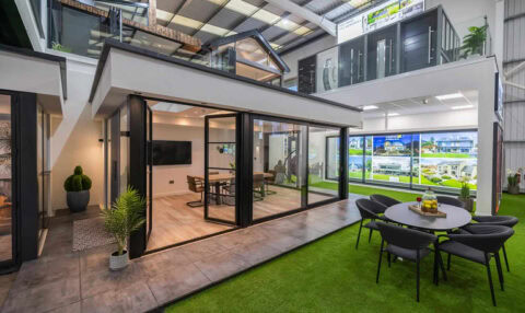Modern indoor showroom featuring a mock house with large bifold doors, dining table, TV, and surrounding artificial grass. Nearby is an outdoor round table with chairs and potted plants beneath a high glass roofing ceiling.