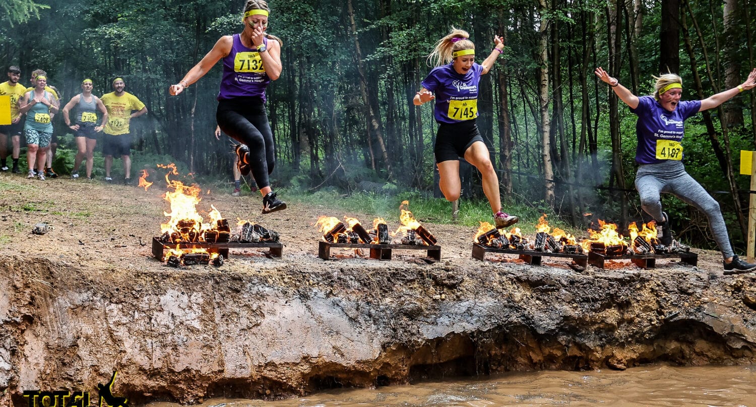 Three women in purple shirts and headbands jump over burning logs during an outdoor obstacle course race, with spectators in yellow shirts watching from behind a row of sliding doors, all set against a backdrop of trees.