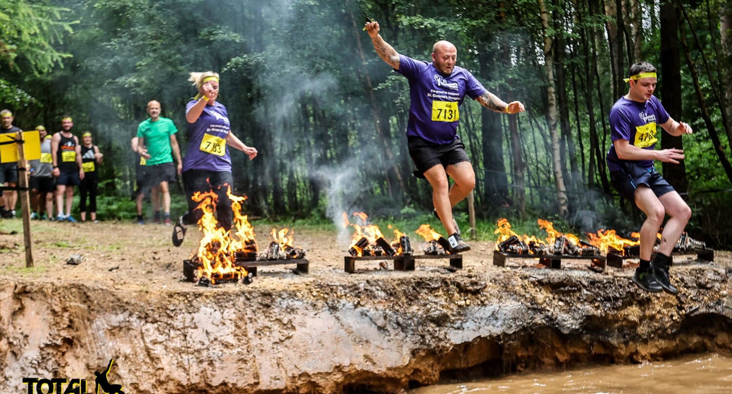 Three athletes in purple shirts and headbands leap over flaming obstacles during an outdoor mud race in a forest. Spectators watch through sliding doors, while the Total Warrior logo appears in the lower left corner.