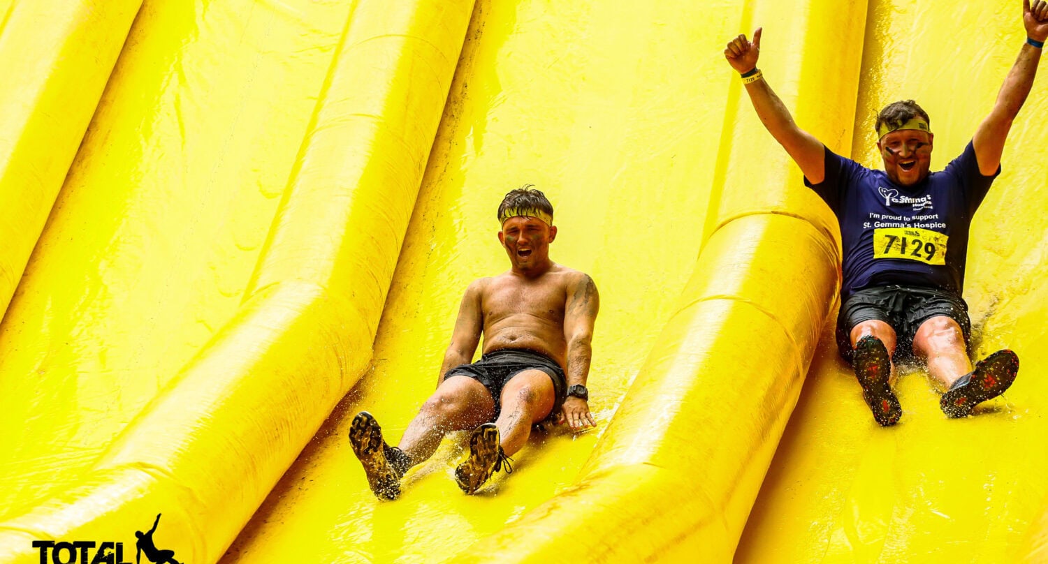 Two people happily slide down a large, bright yellow inflatable slide at a Total Warrior event, both smiling and raising their arms in excitement. The logo Total Warrior appears in the bottom left corner, much like the entryway of stylish front doors.