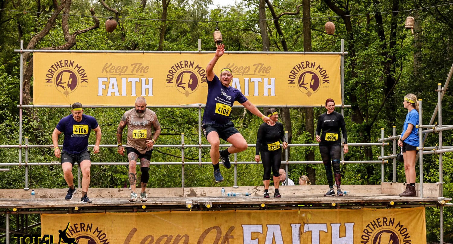 Six participants leap off a wooden platform labeled “Leap of Faith” during an outdoor obstacle event, surrounded by trees, event banners, and a canopy featuring bespoke glass solutions, with some midair and others preparing to jump.