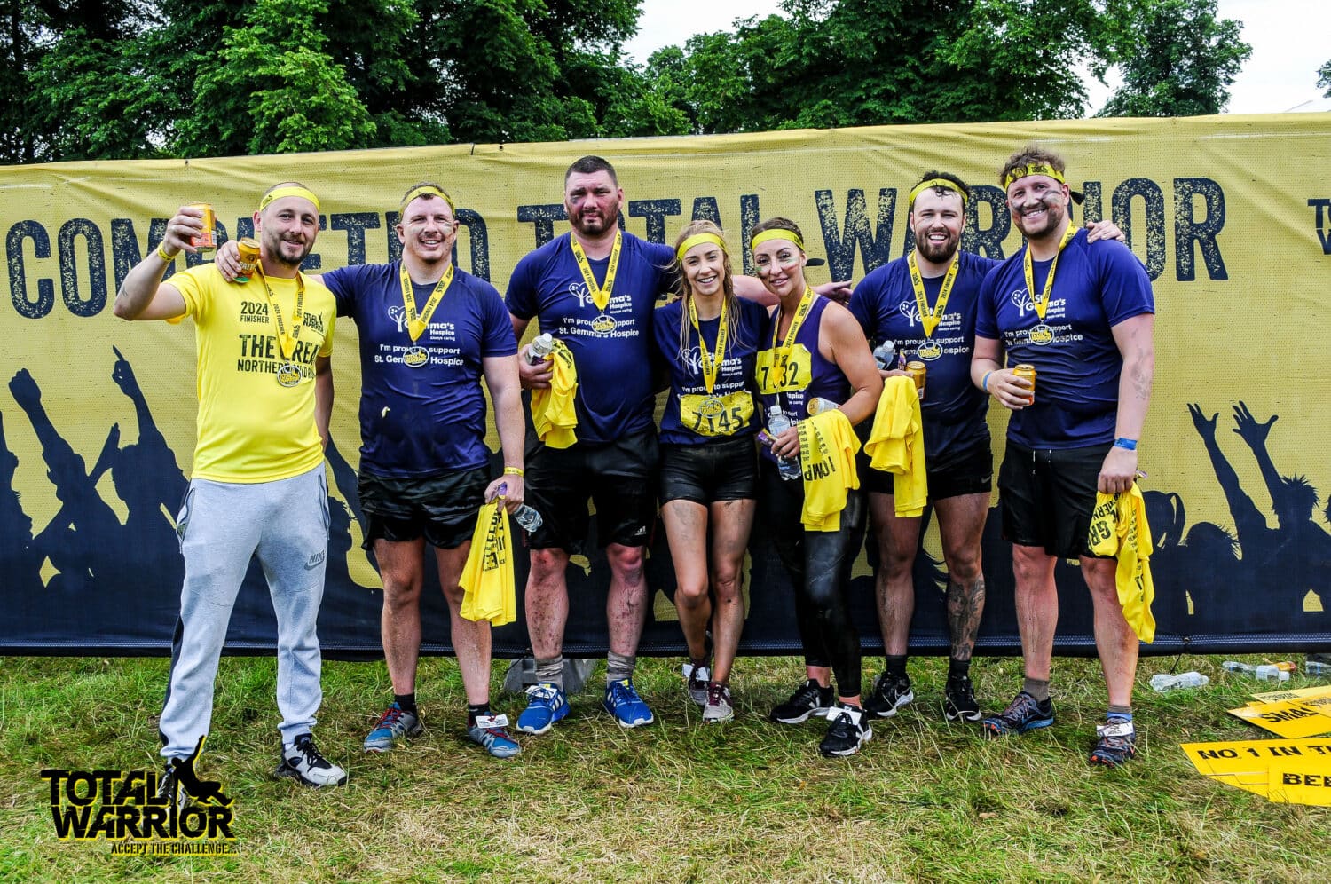 A group of seven muddy people wearing matching “The Gym” shirts and yellow medals stand smiling together after a race, holding yellow towels in front of a Total Warrior event banner outdoors beneath glass roofing.