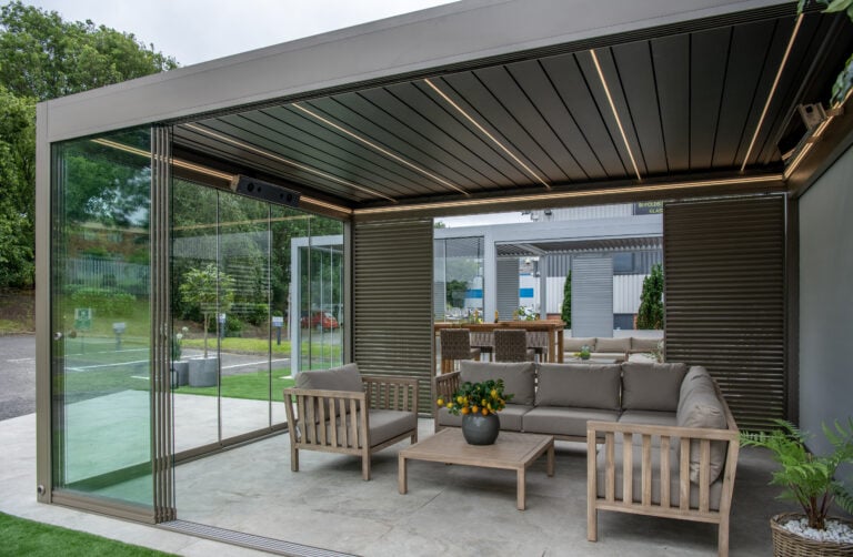 Modern outdoor patio with a covered seating area, featuring bespoke glass solutions like sliding doors, wooden furniture with gray cushions, a coffee table with a plant, and a view of another seating area and greenery outside.