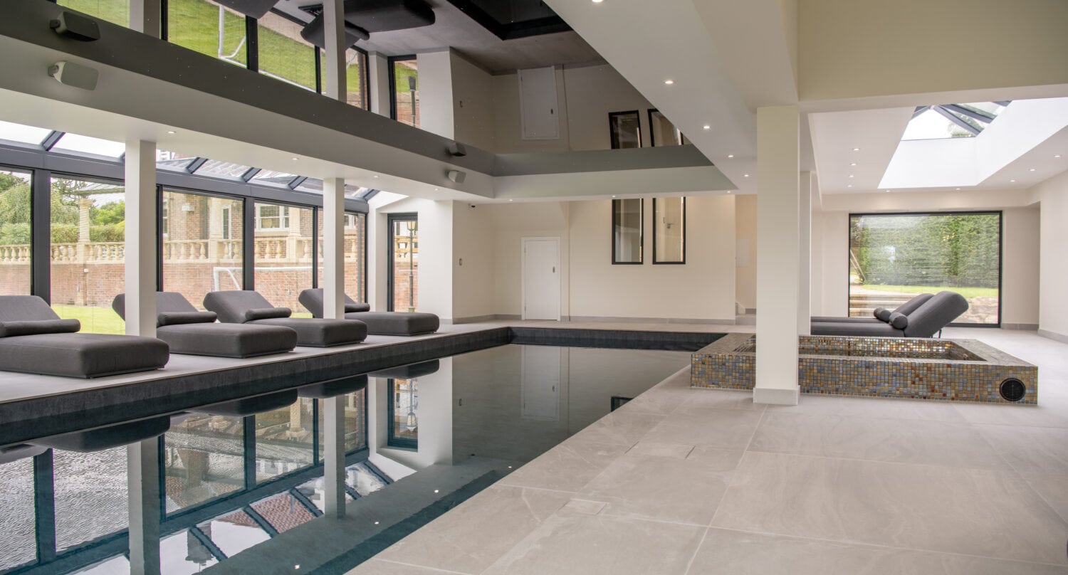 Modern indoor swimming pool area with lounge chairs along the sides, large windows and glass roofing letting in natural light, and a hot tub in the corner. The room features light-colored tiles and a clean, spacious design.