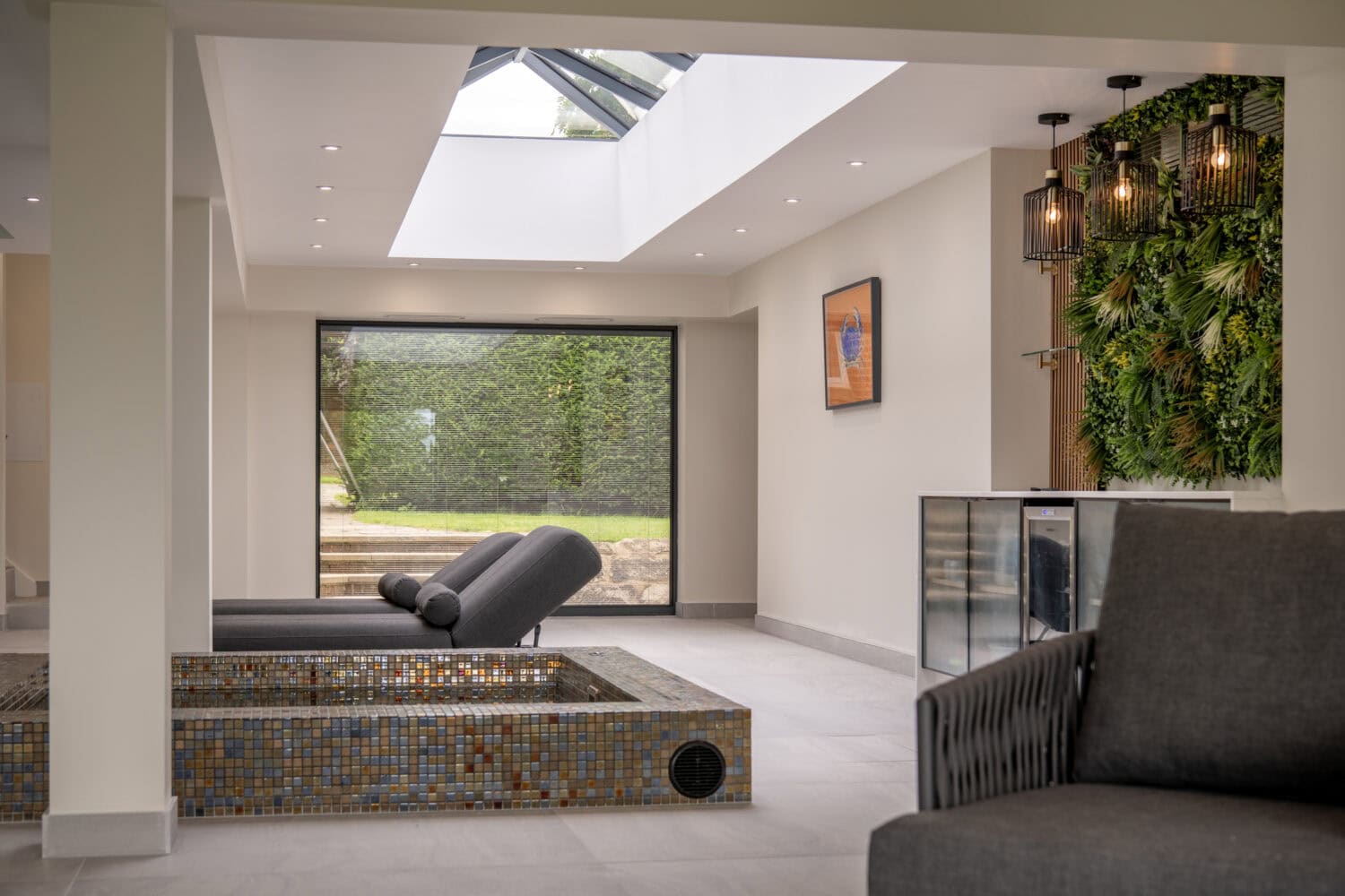 Modern indoor lounge with two gray chaise lounges by a tiled, empty sunken tub, large window, vertical garden wall, glass ceiling, and contemporary hanging lights. Bifold doors invite natural light into the minimalist space.