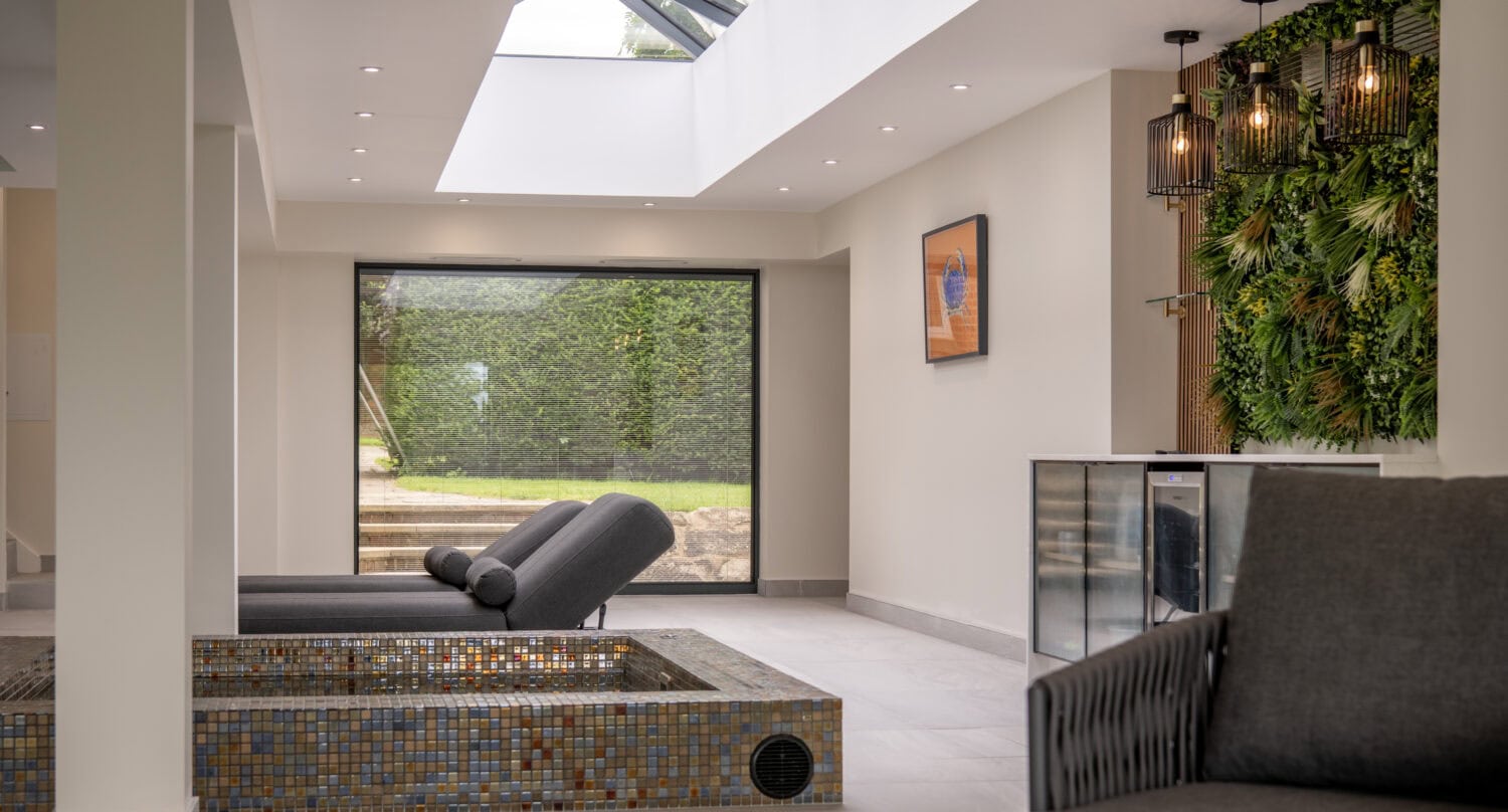 Modern indoor lounge with two gray chaise lounges by a tiled, empty sunken tub, large window, vertical garden wall, glass ceiling, and contemporary hanging lights. Bifold doors invite natural light into the minimalist space.