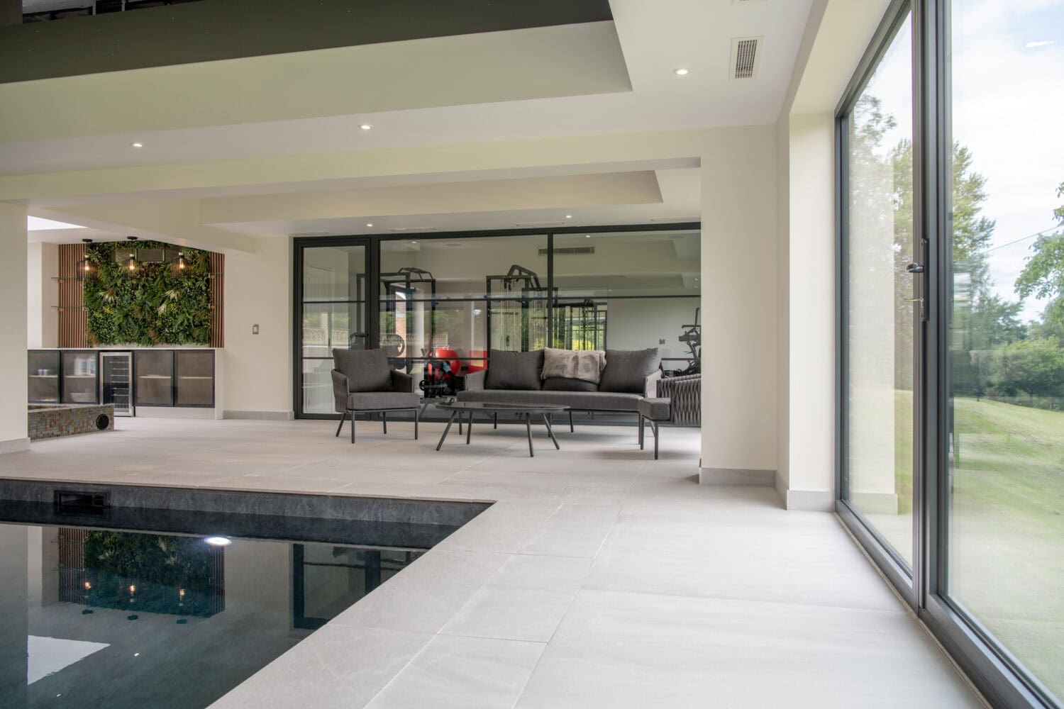 Modern indoor space with large glass windows and sliding doors, a dark-edged pool, light-colored tiled floor, and contemporary seating area with sofas. Greenery is visible outside and on an indoor plant wall.