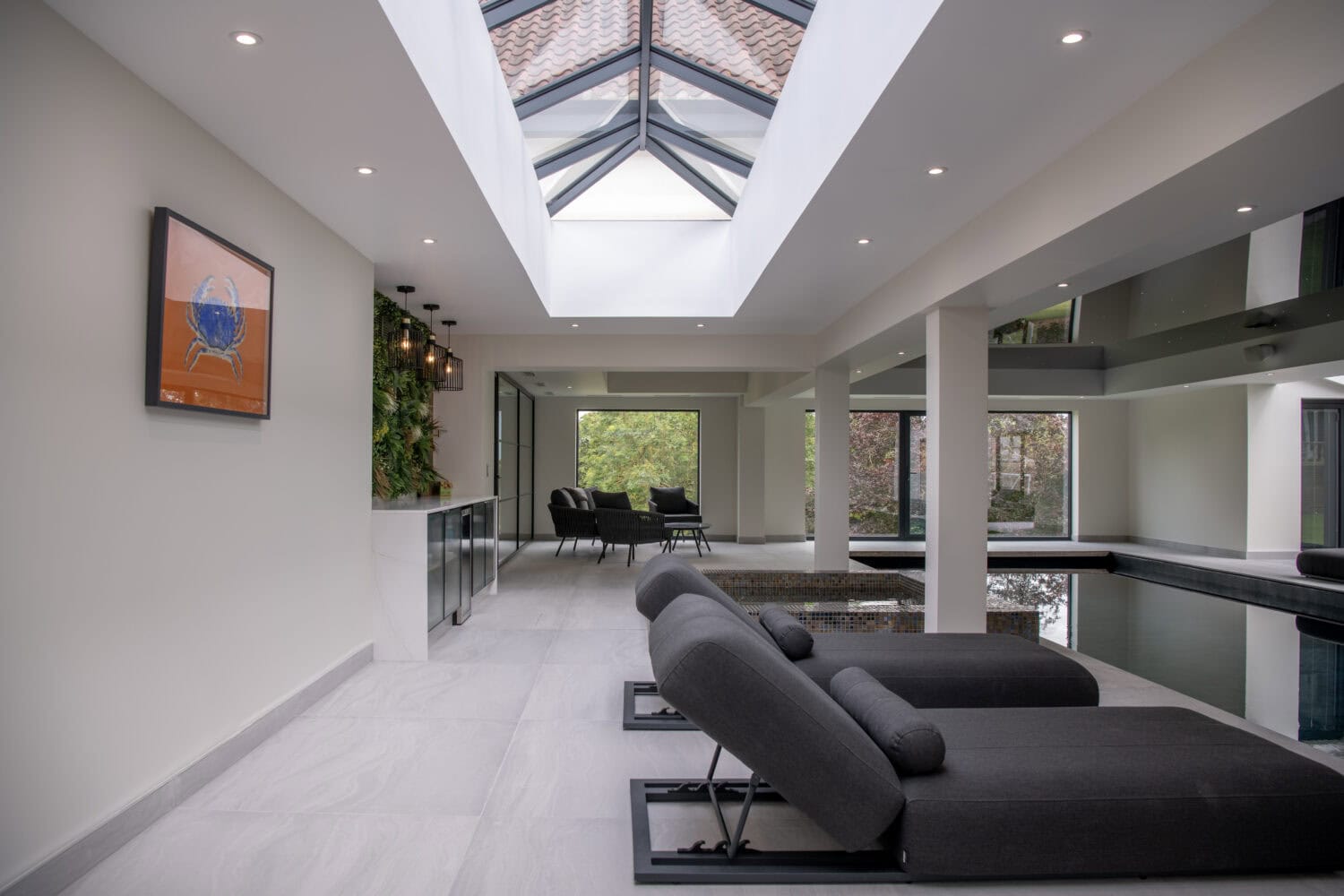 Modern indoor pool area with two gray lounge chairs, a skylight ceiling, wall art, greenery, and large windows created with bespoke glass solutions overlooking trees; a dining area is visible in the background. The space features sleek, minimalistic design.