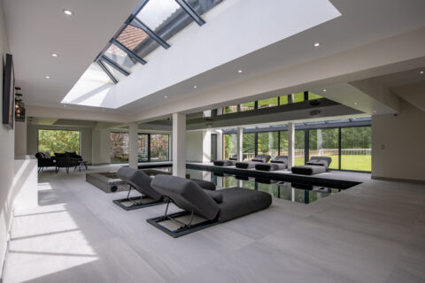 Modern indoor pool area with several black lounge chairs, large windows, skylights, and bespoke glass solutions offering a seamless view of the green garden outside. The bright, spacious space features sleek, contemporary design elements.