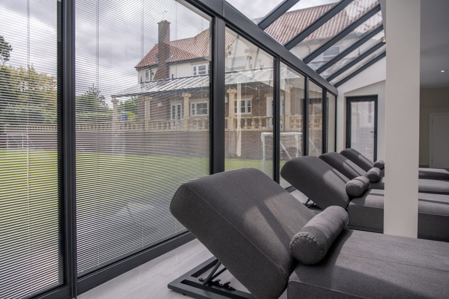 Modern relaxation area with gray recliners facing large bifold doors and glass windows, overlooking a green lawn and a brick house outside. Blinds partially cover the windows, letting in natural light.