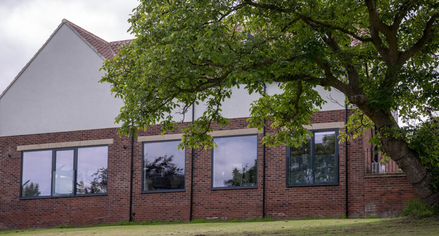 A brick building with large windows and bespoke glass solutions sits behind a grassy lawn and a leafy tree, under a cloudy sky.
