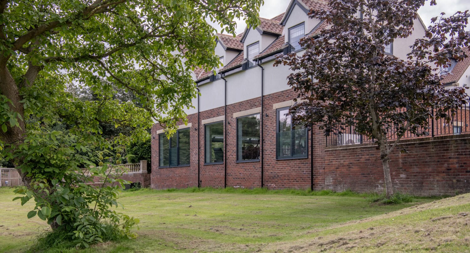 A modern red-brick building with large windows, white upper walls, and sleek bifold doors stands beside a grassy lawn with green trees. The sky is partly cloudy.